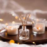 Home perfume in glass bottle with wood sticks, scented burn candles  tray in bedroom close up. Aromatherapy cozy atmosphere lifestyle. Winter warm xmas season.